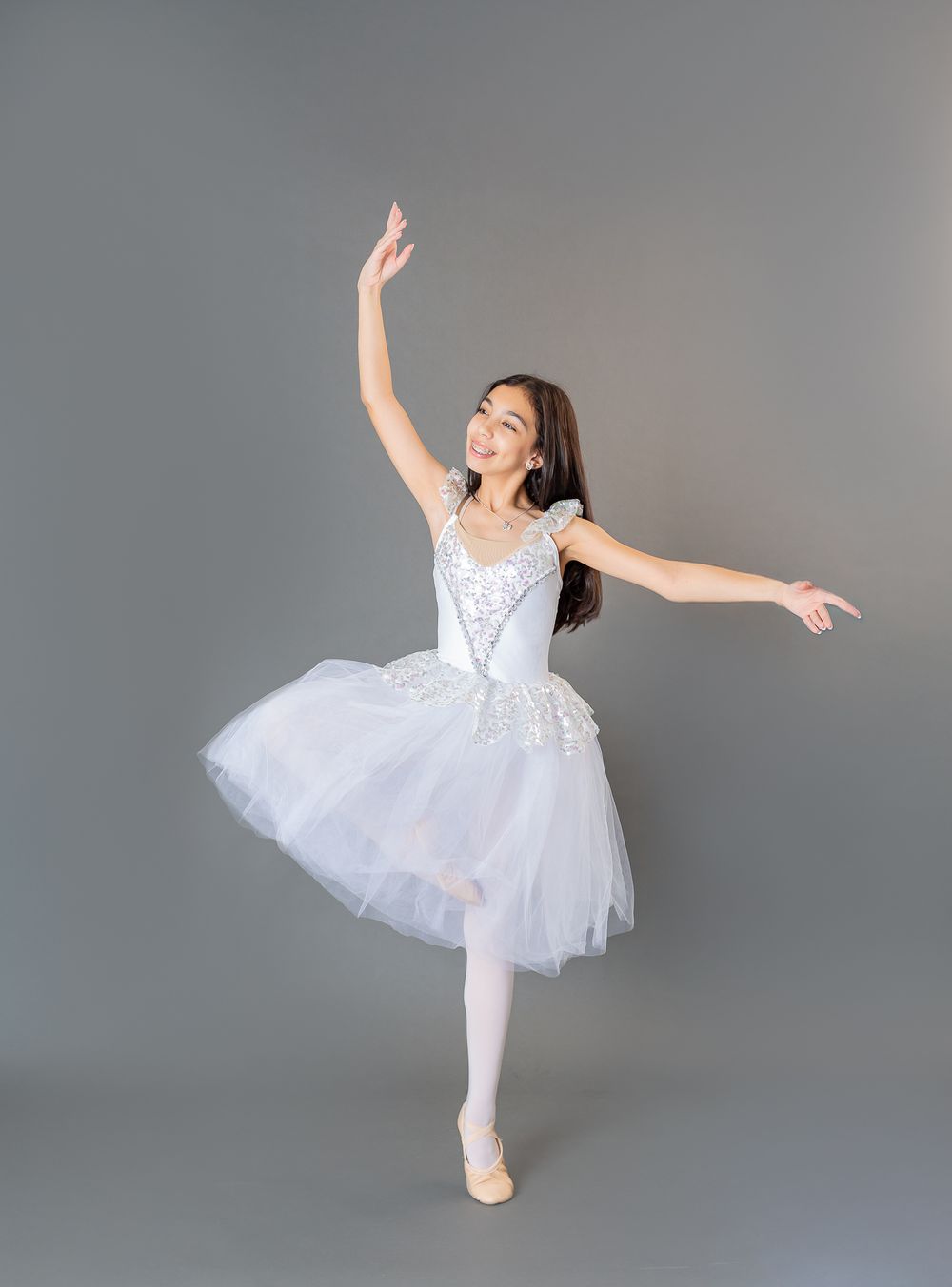 A dancer in a white tutu performs an elegant arabesque pose against a gray studio backdrop.