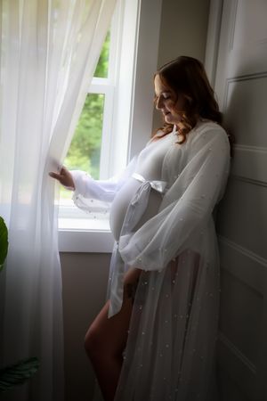 Dreamy maternity photo by window with flowing white sheer curtains and natural light.