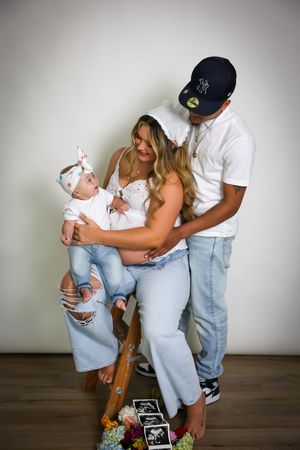 A family poses together holding a baby while surrounded by ultrasound photos and decorative items on a wooden floor.