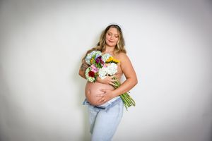 Expectant mother in jeans holds colorful bouquet while showing off baby bump against white wall.