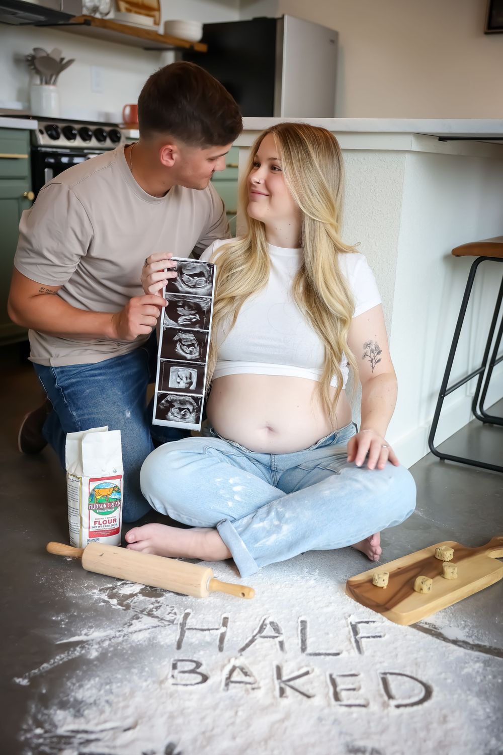 Creative pregnancy announcement with 'Half Baked' written in flour on kitchen counter.