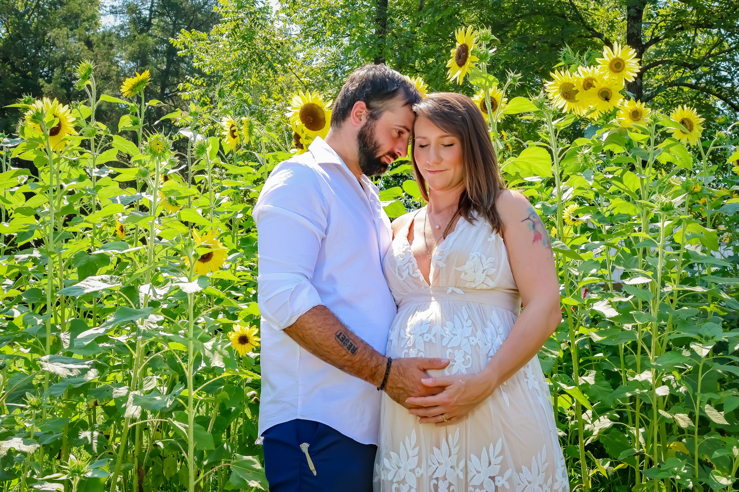 A maternity photo in a field of bright yellow sunflowers on a sunny day.
