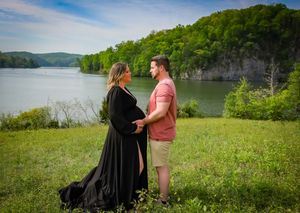 Maternity photoshoot in black dress against scenic lake and mountain backdrop.