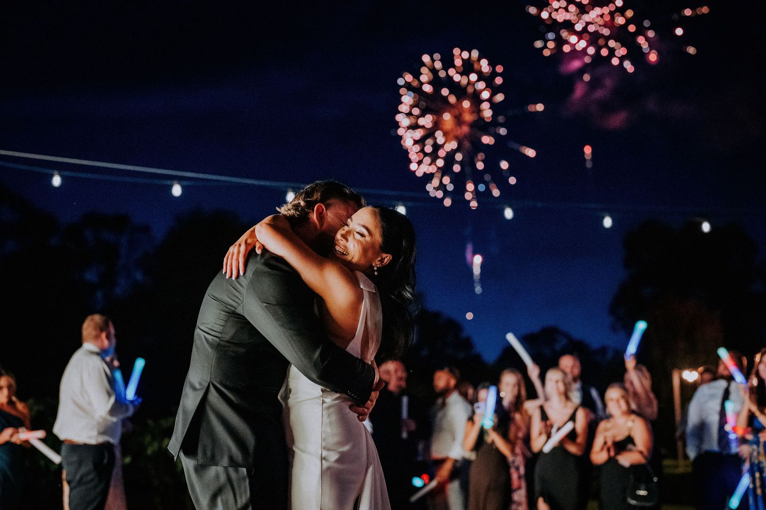 Wedding guests dance under twinkling lights and fireworks during an outdoor evening reception celebration.