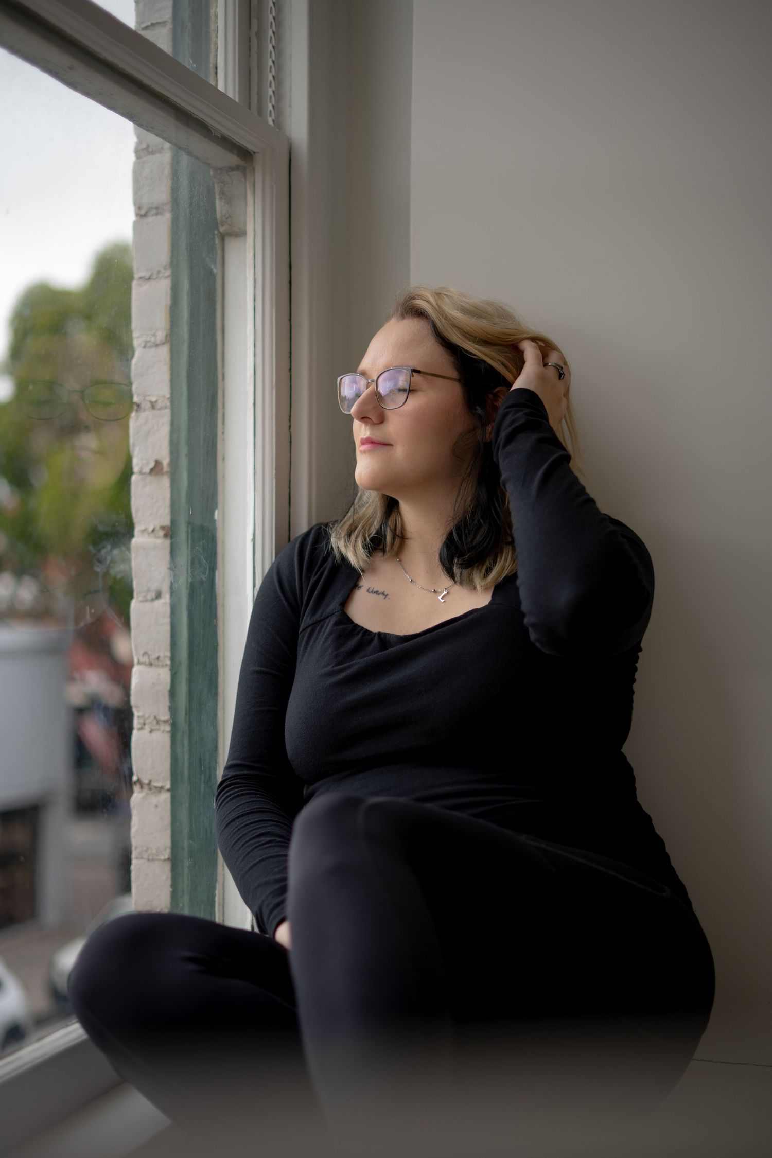 Person in glasses wearing black sits contemplatively by a window in natural light.