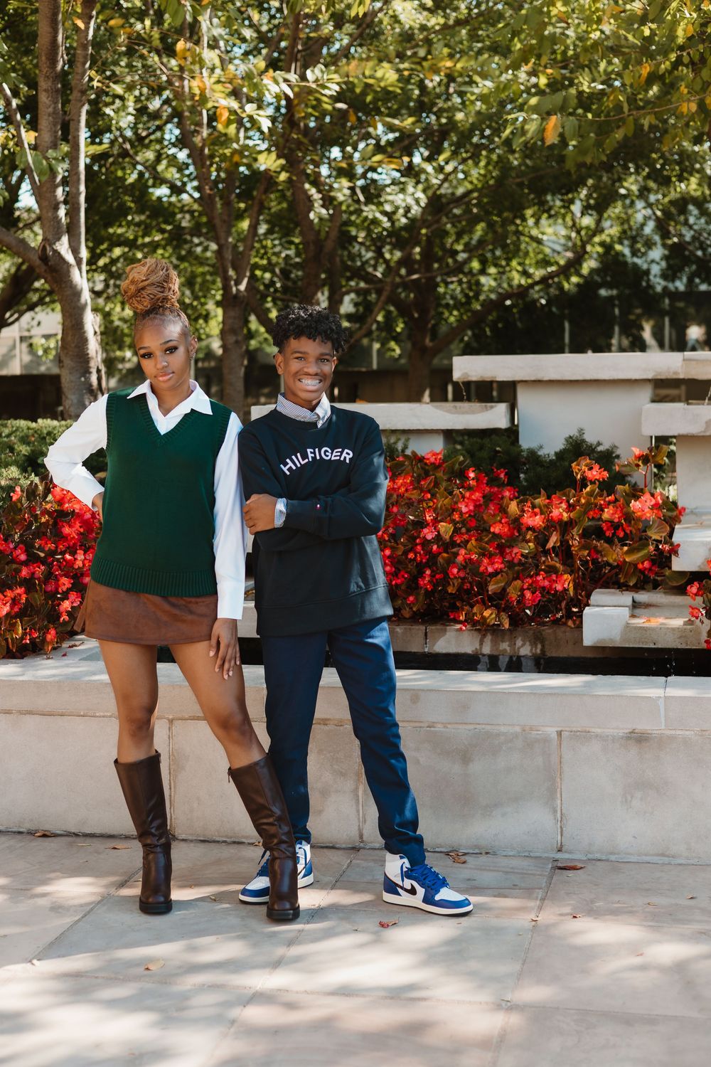 Two young people in casual fall outfits pose together outdoors near red flowers and green trees.