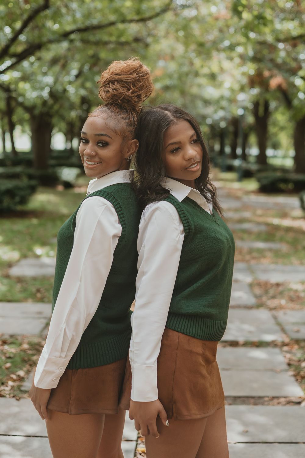 Two people in coordinating green sweaters and brown skirts stand back to back on a sidewalk in autumn.