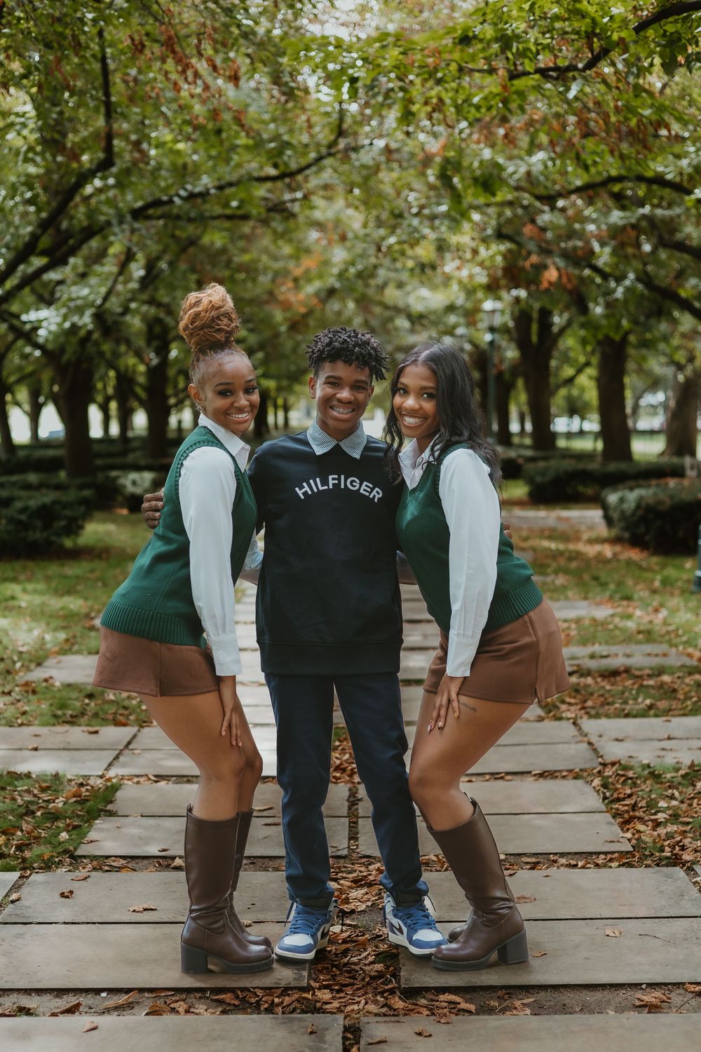 Three friends pose together on a stone path in a park during autumn wearing coordinated outfits with boots and sweaters.