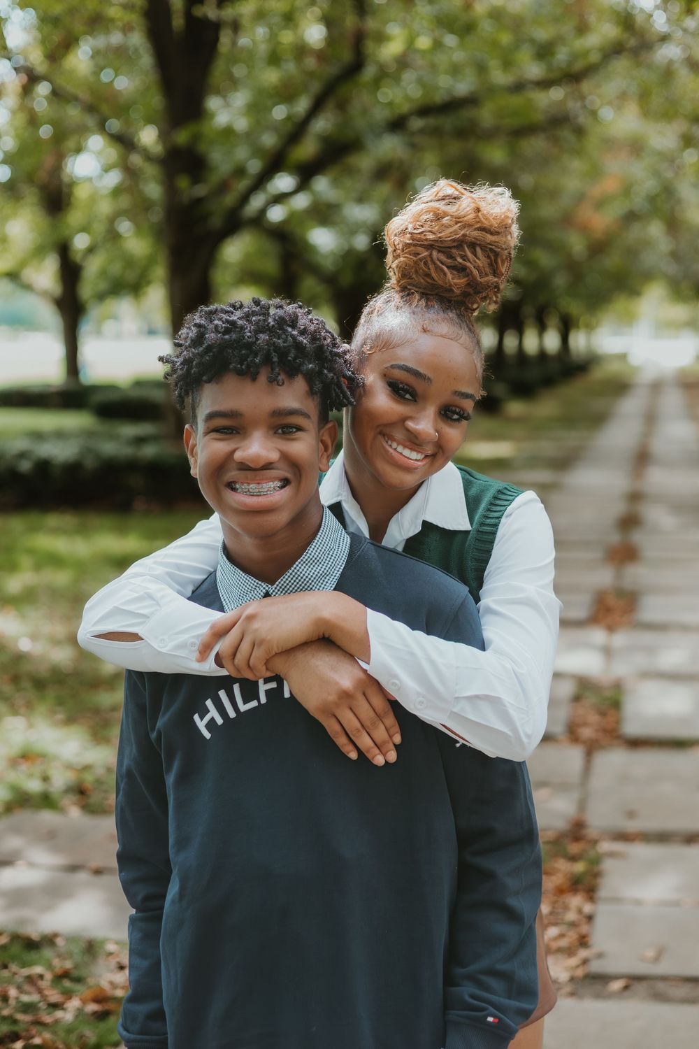 Two people share a joyful embrace while wearing casual clothing outdoors on an autumn day with fallen leaves visible.