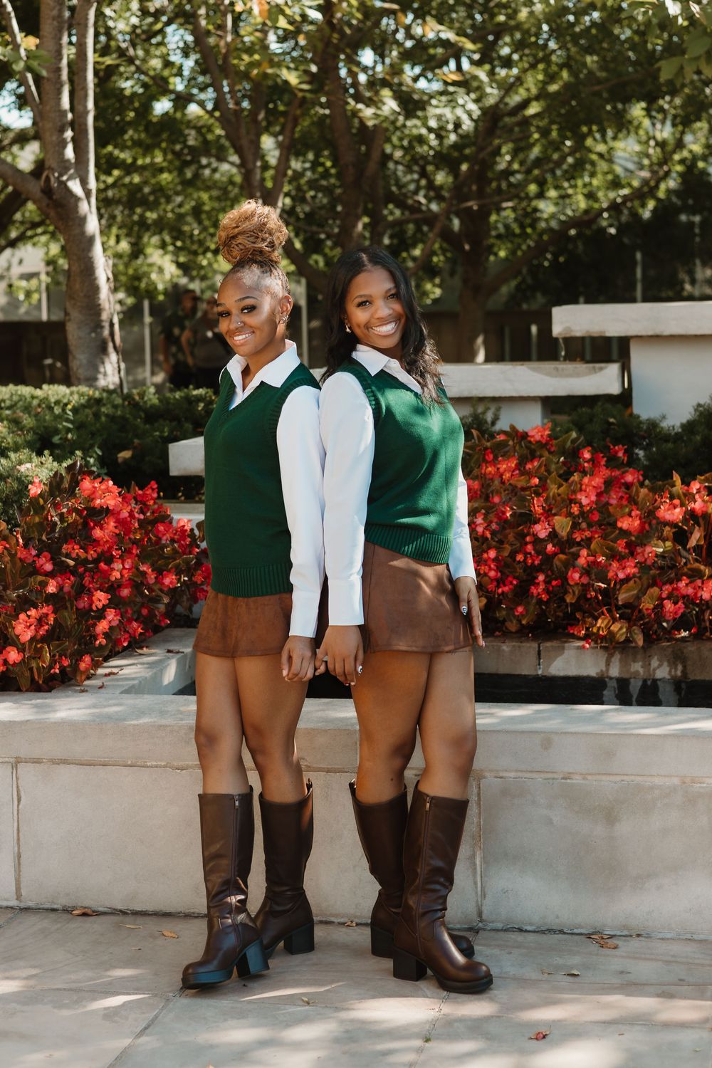 Two people wearing brown lederhosen shorts and boots stand in front of red flowers and greenery in an outdoor setting.