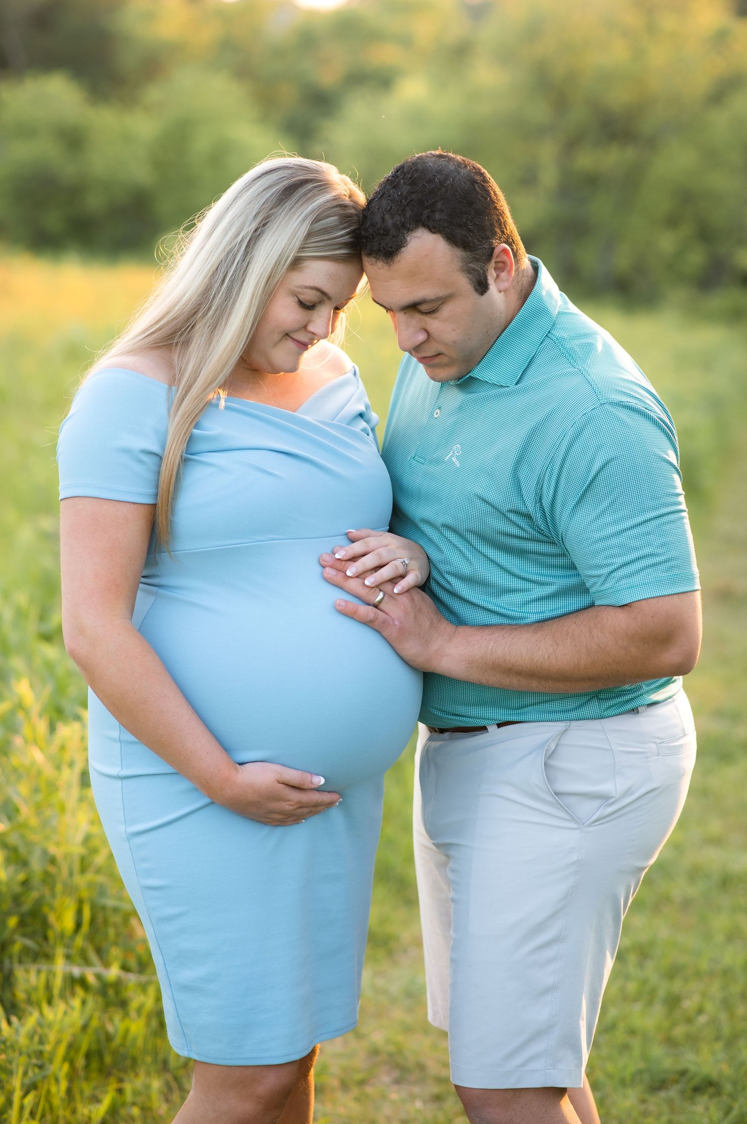 Expectant couple shares tender moments in a sunlit meadow wearing coordinating blue and white outfits.