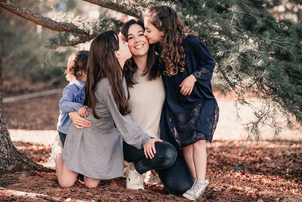 Three people share a tender moment outdoors on fallen autumn leaves under a tree.