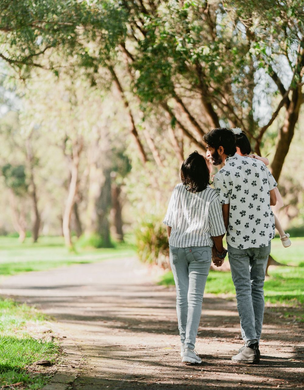 Two people walking together on a tree-lined dirt path in a park on a sunny day.