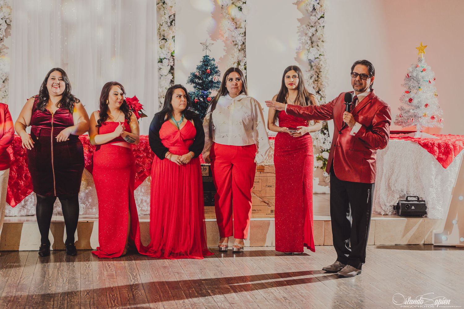 A group of singers in coordinating red and white outfits perform in front of a decorated holiday backdrop with Christmas trees.