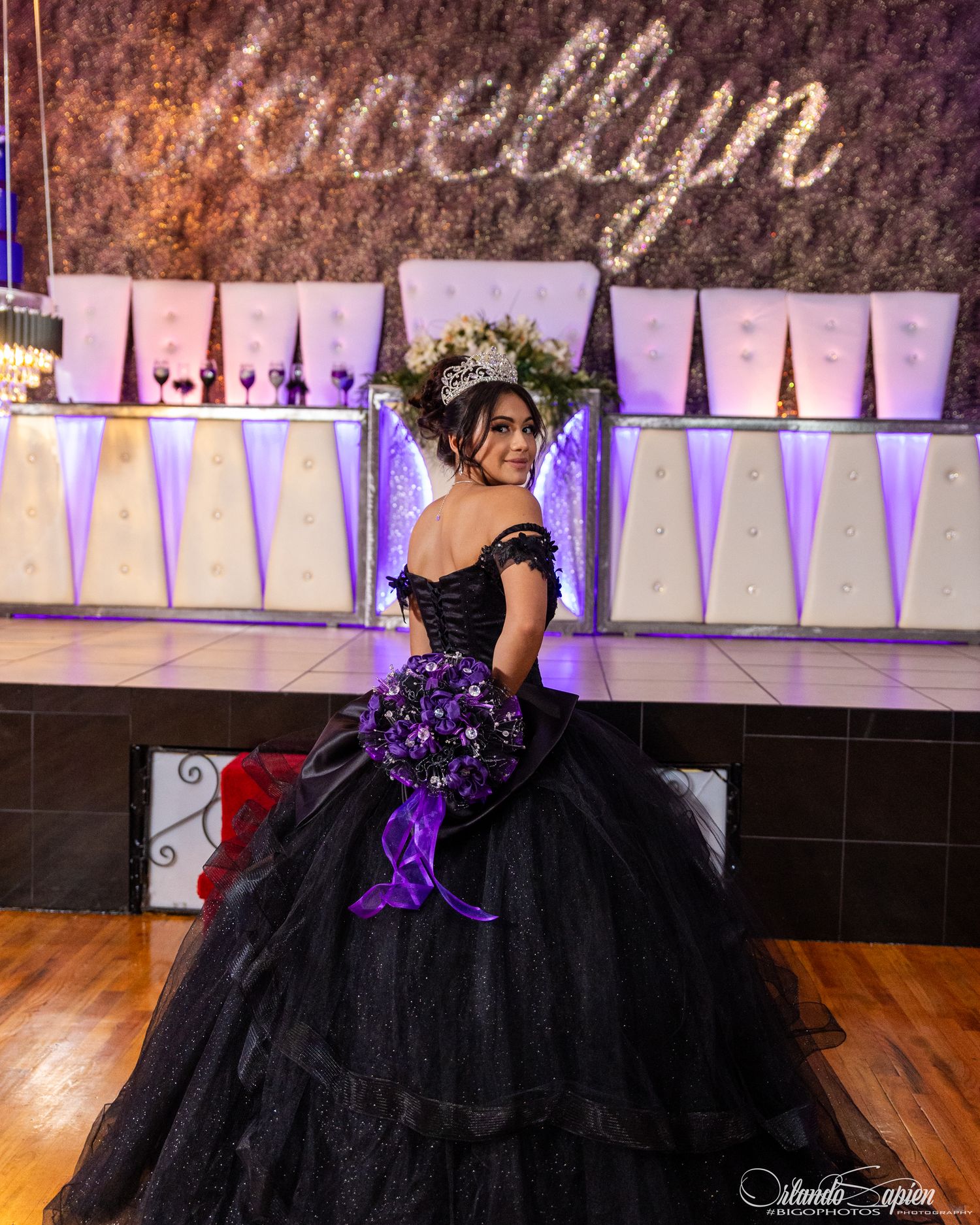 A dramatic black ball gown with purple accents and tulle layers sits elegantly on a polished wooden dance floor.