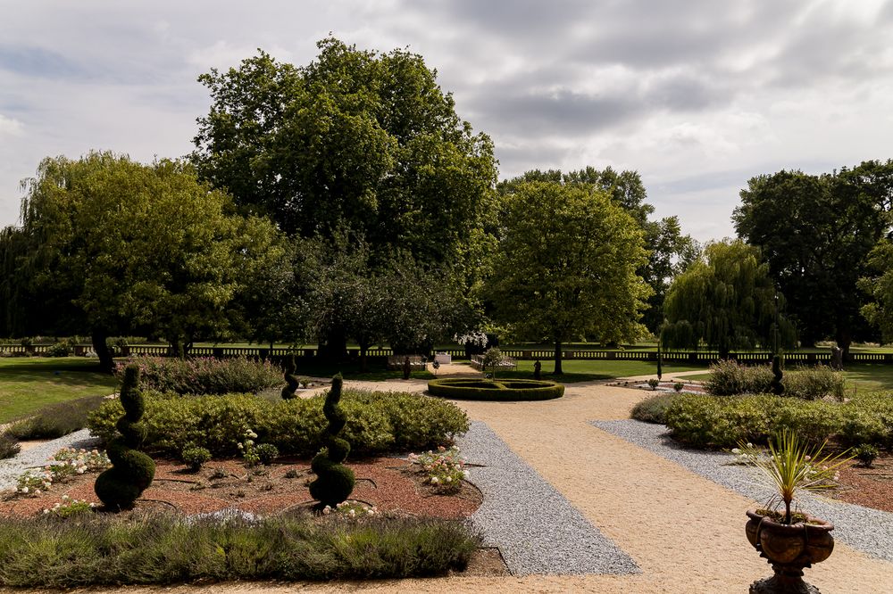 A landscaped garden path with gravel walkways and manicured flower beds under cloudy skies.