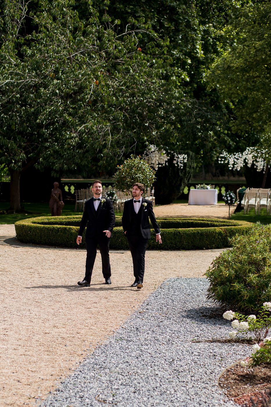 Two people in formal attire walking along a gravel garden path bordered by manicured hedges.