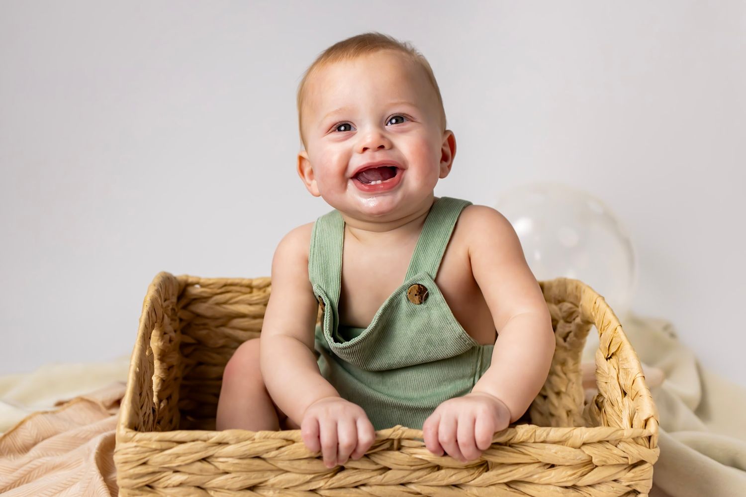 Baby wearing olive green tank top sits in woven basket smiling with joy and enthusiasm.
