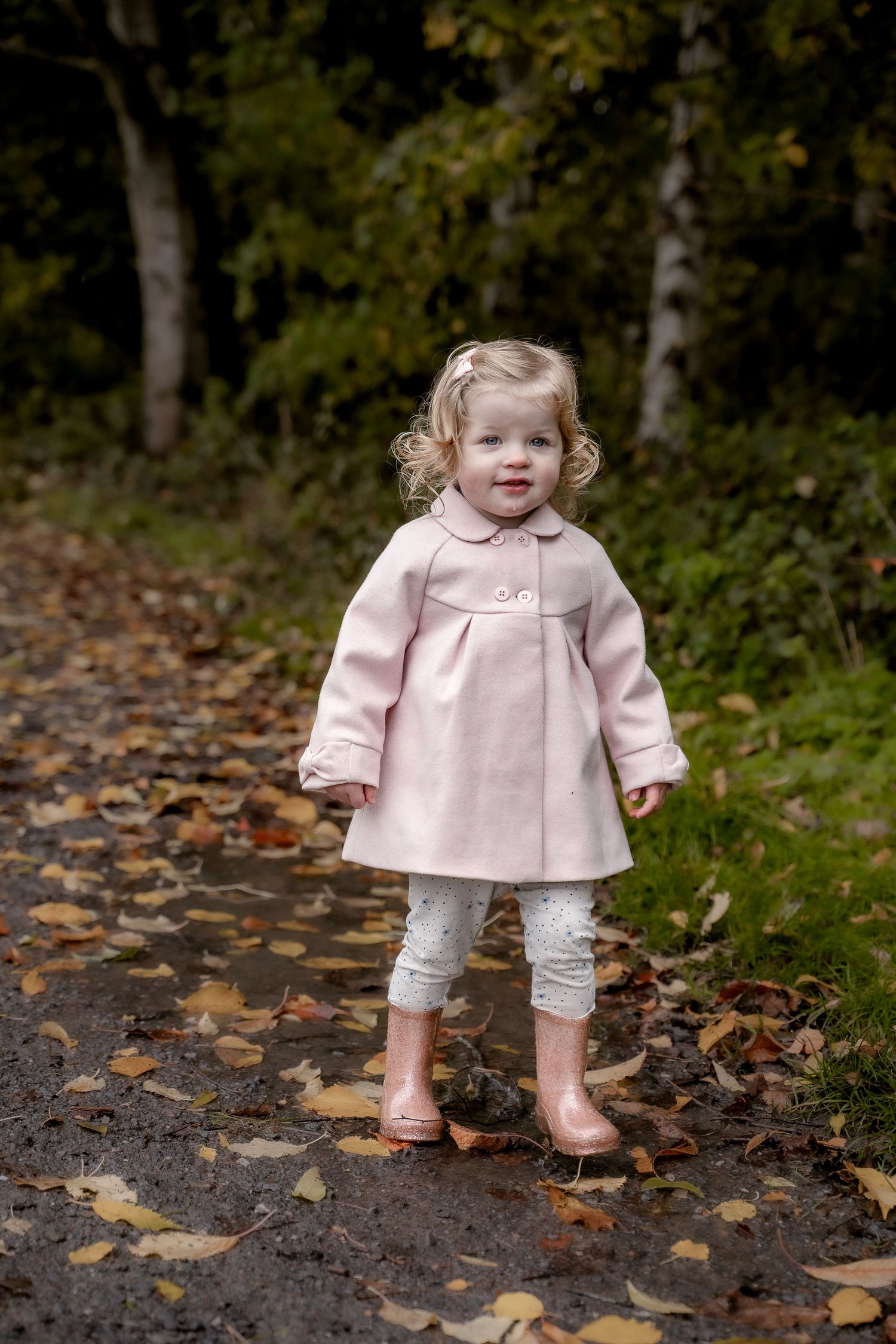 A small child wearing a pink coat and white tights stands on a path covered in autumn leaves.