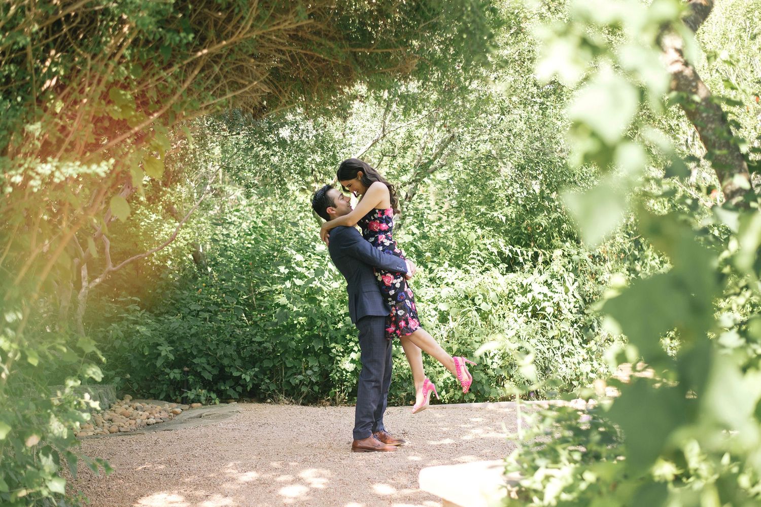 A romantic couple embraces on a sunlit garden path surrounded by lush greenery.