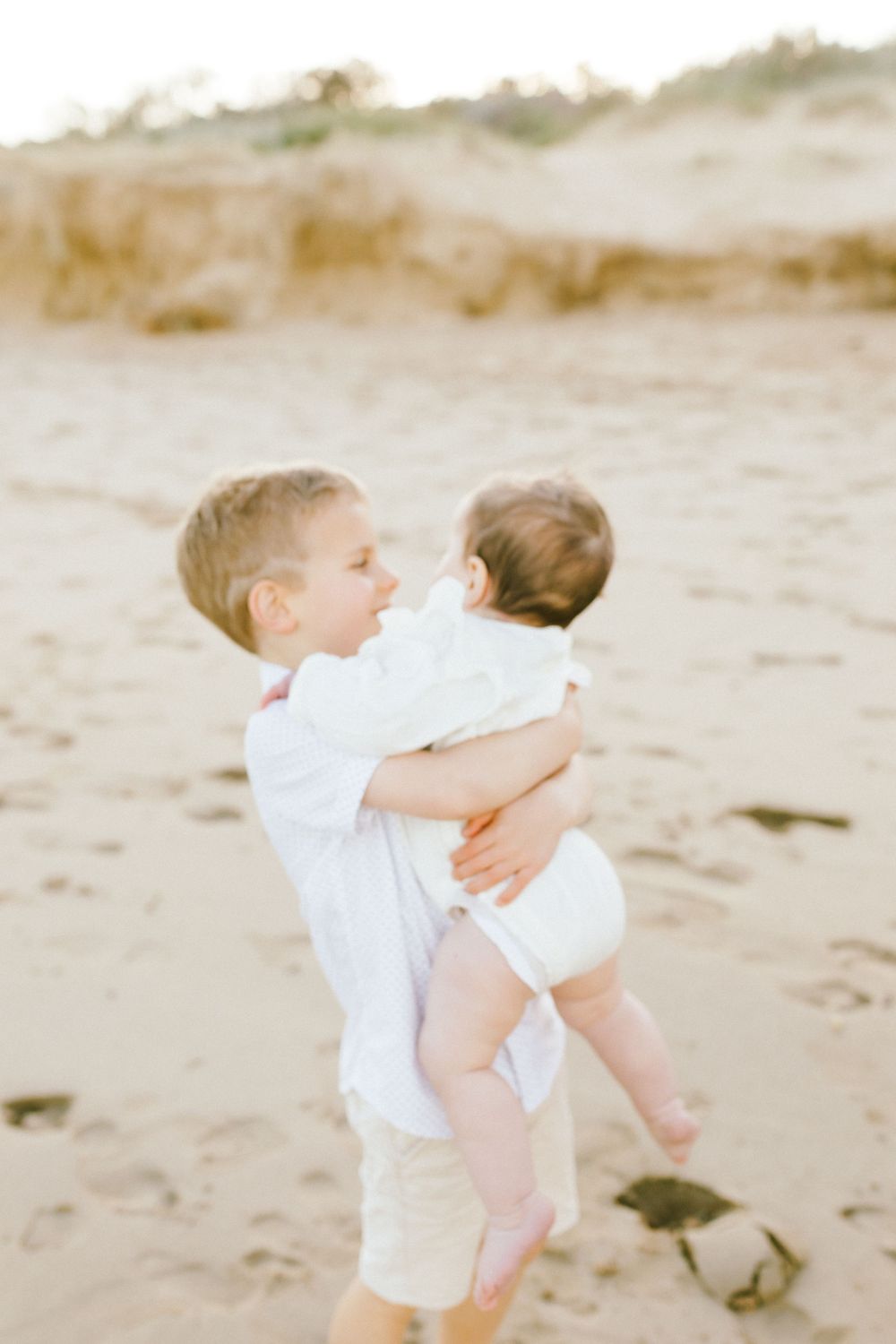Two young children in white outfits embrace while playing together on a sandy beach during golden hour.