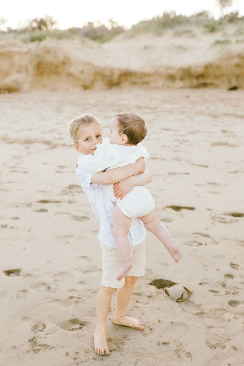 Two children in white outfits play together on a sandy beach during a sunny day.