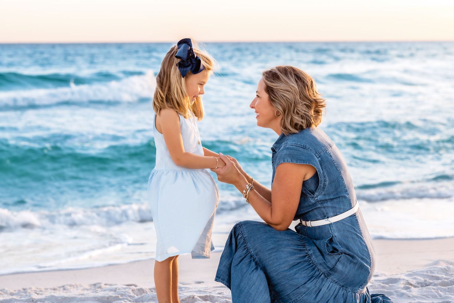 Two people embrace while sitting on a sandy beach with ocean waves in the background.