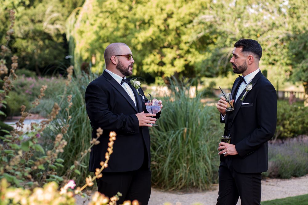 Two men in formal attire having drinks and conversation in a garden setting at sunset.