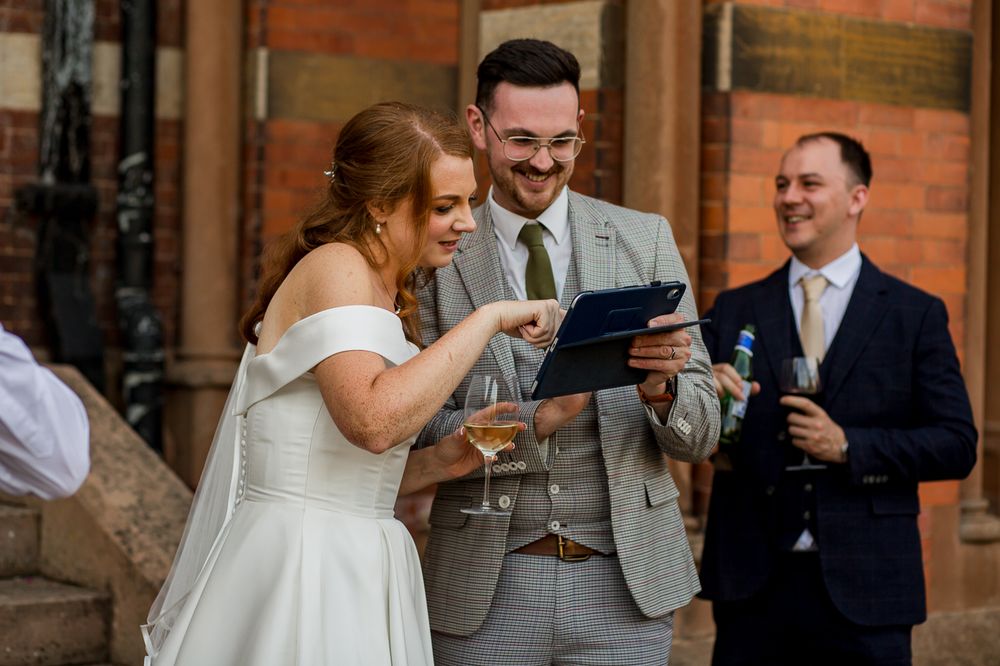 Wedding couple and guest looking at digital device outside brick building.