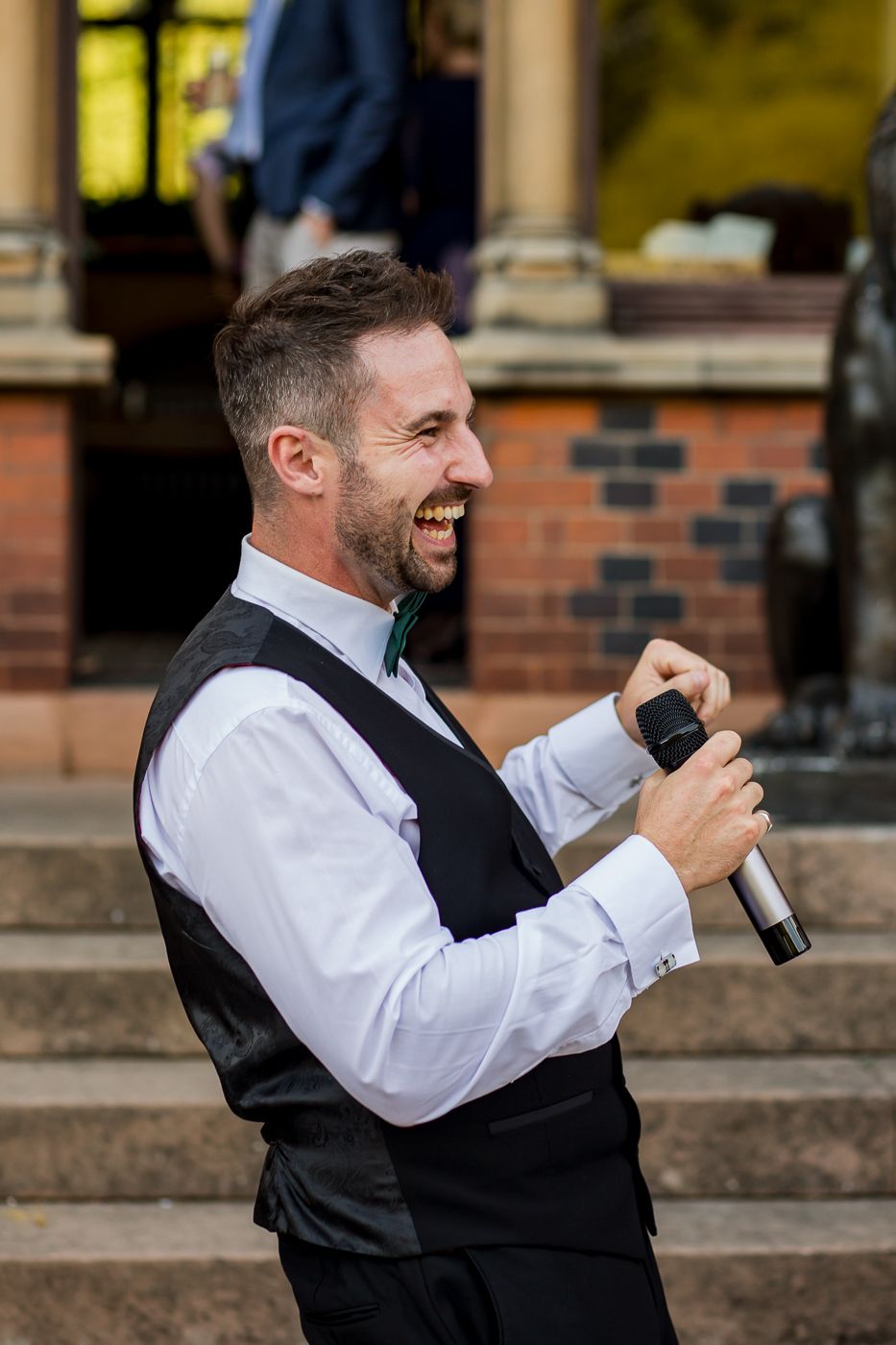 Wedding entertainer in vest and white shirt performing with microphone on stone steps.
