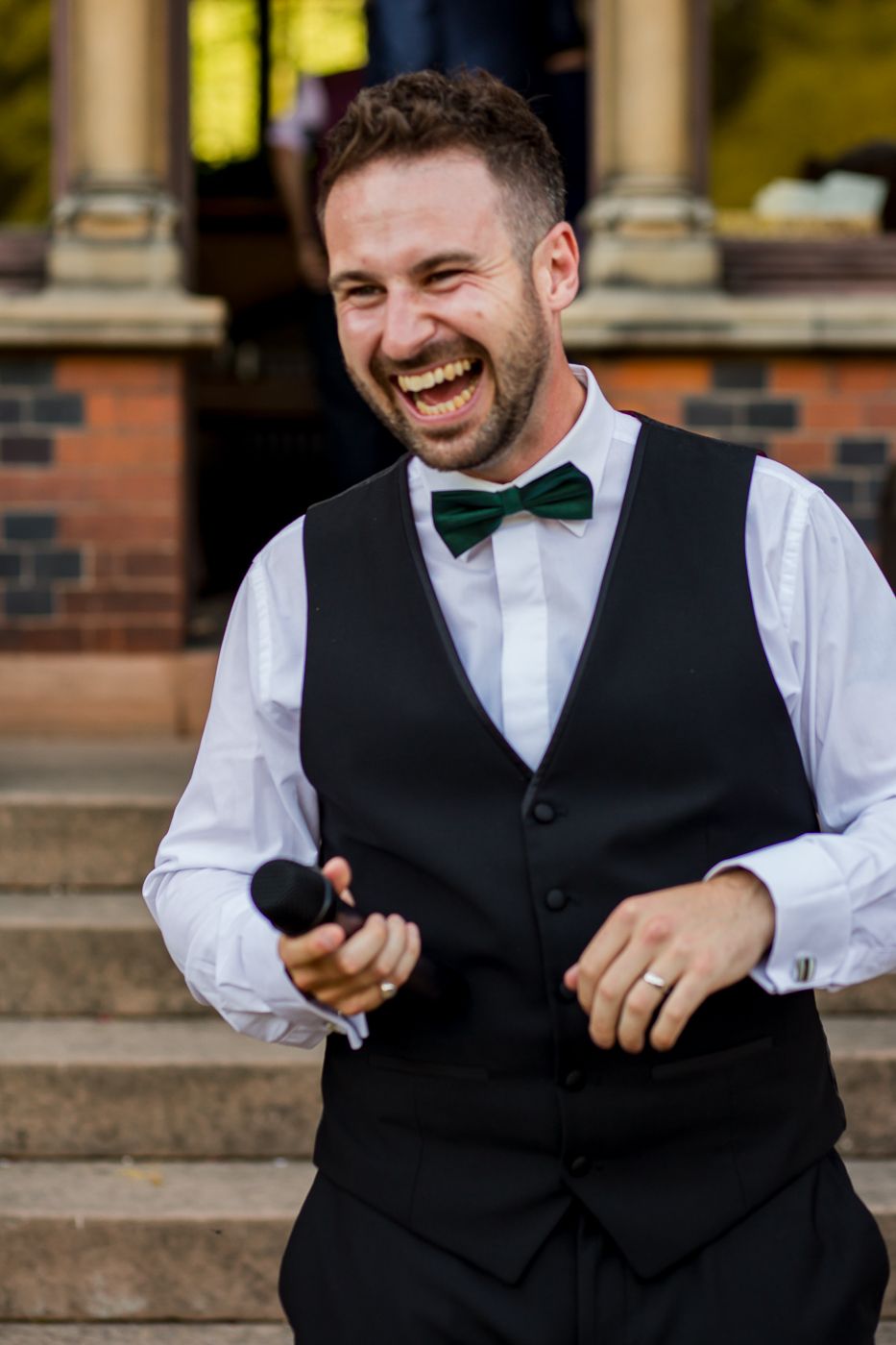 Entertainer in bow tie and vest laughing while holding microphone during outdoor performance.