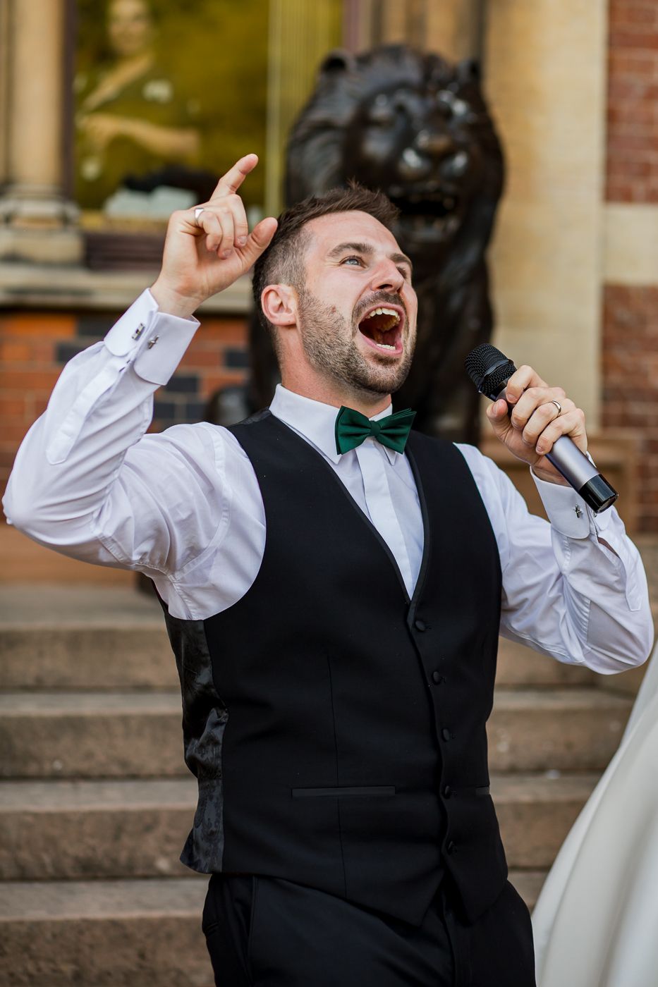 Animated performer in bow tie gestures upward while singing at outdoor wedding venue.