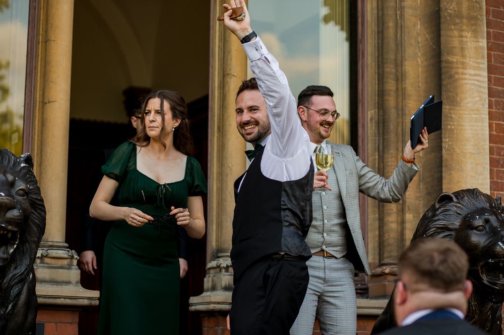 Wedding party members laugh and socialize outside a historic brick venue.