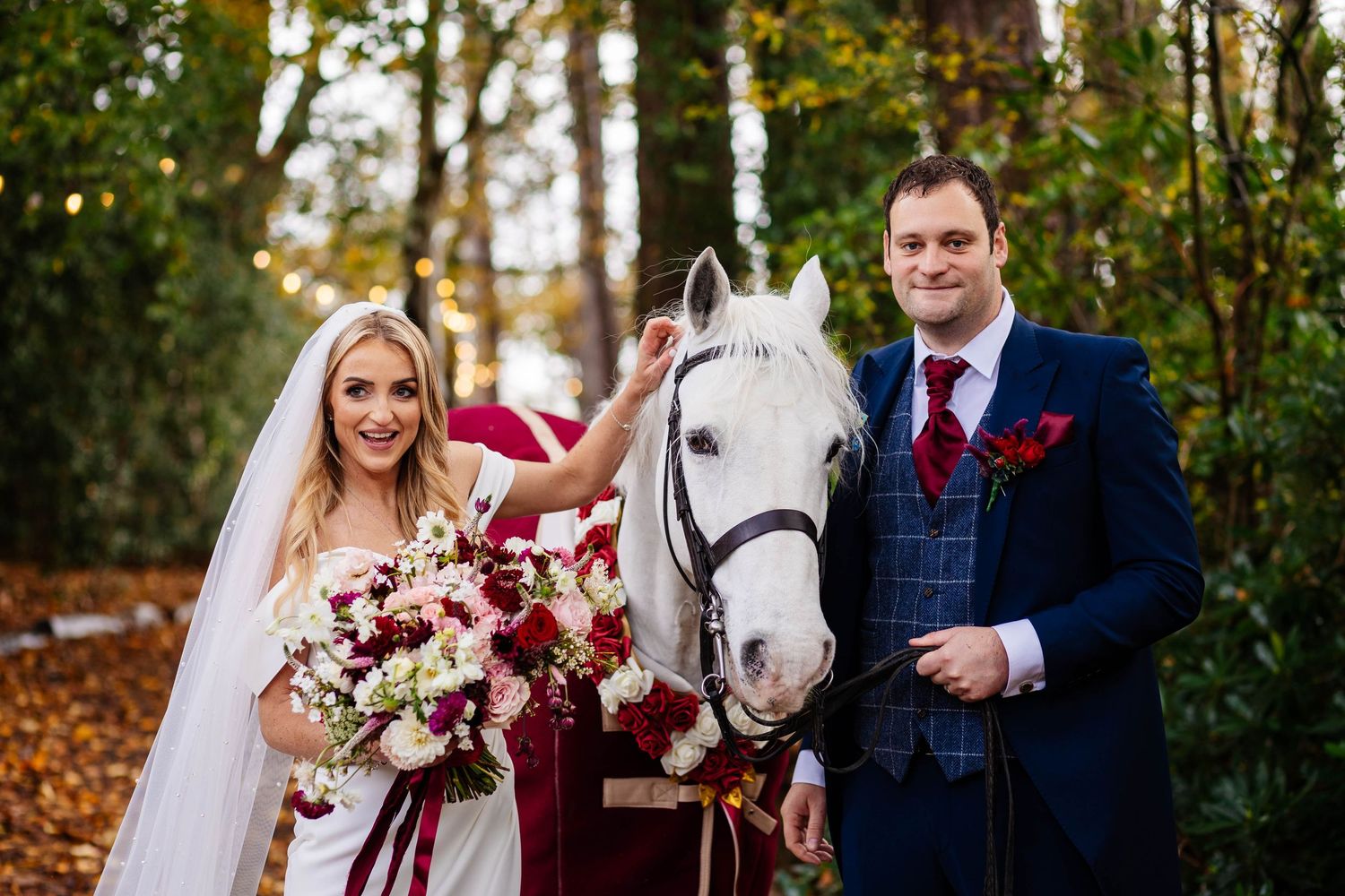 A couple poses with a white horse during their wedding in a wooded setting at Hobbit Hill, captured by Lancashire wedding photographer