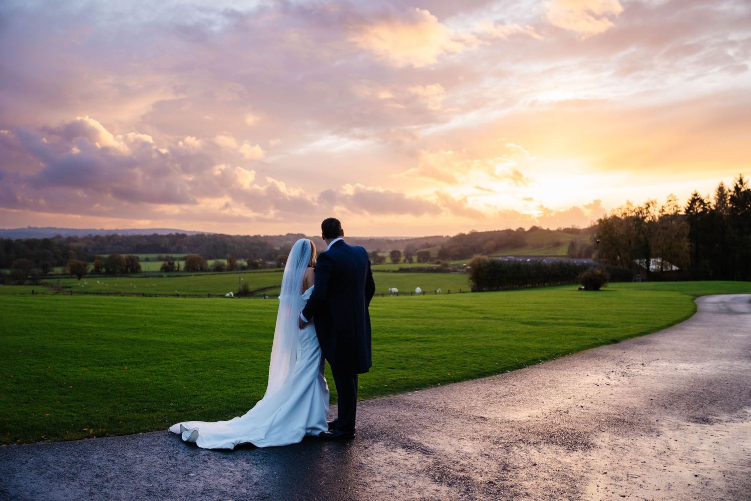 A couple stands against a stunning sunset over rolling countryside hills and meadows at Hobbit Hill captured by Lynette Matthews Photography