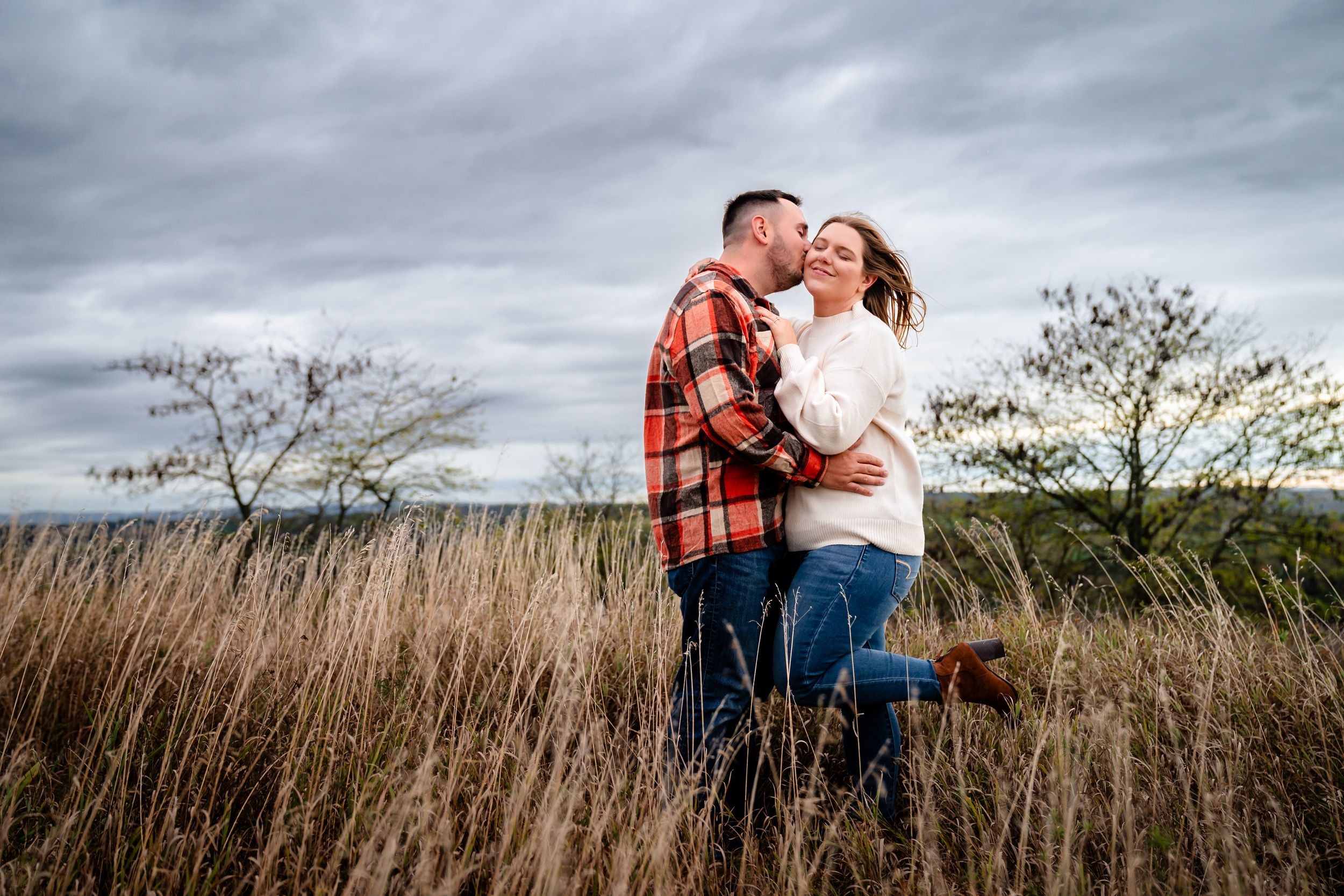 A couple embraces in a field of golden grass with dramatic cloudy skies overhead.