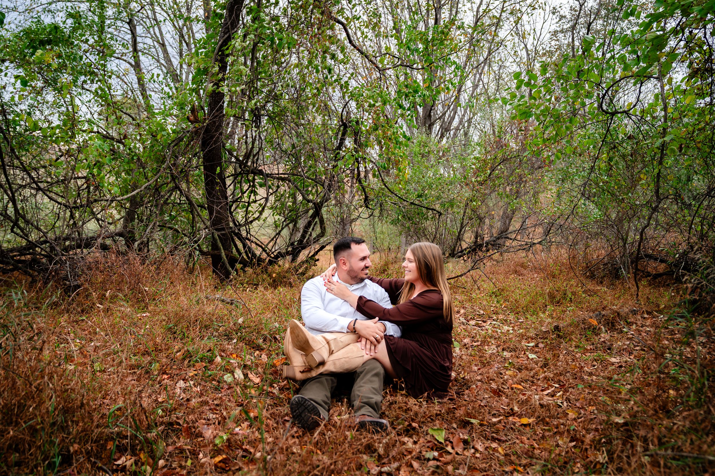 Couple sitting together on fallen leaves during an autumn engagement photoshoot.