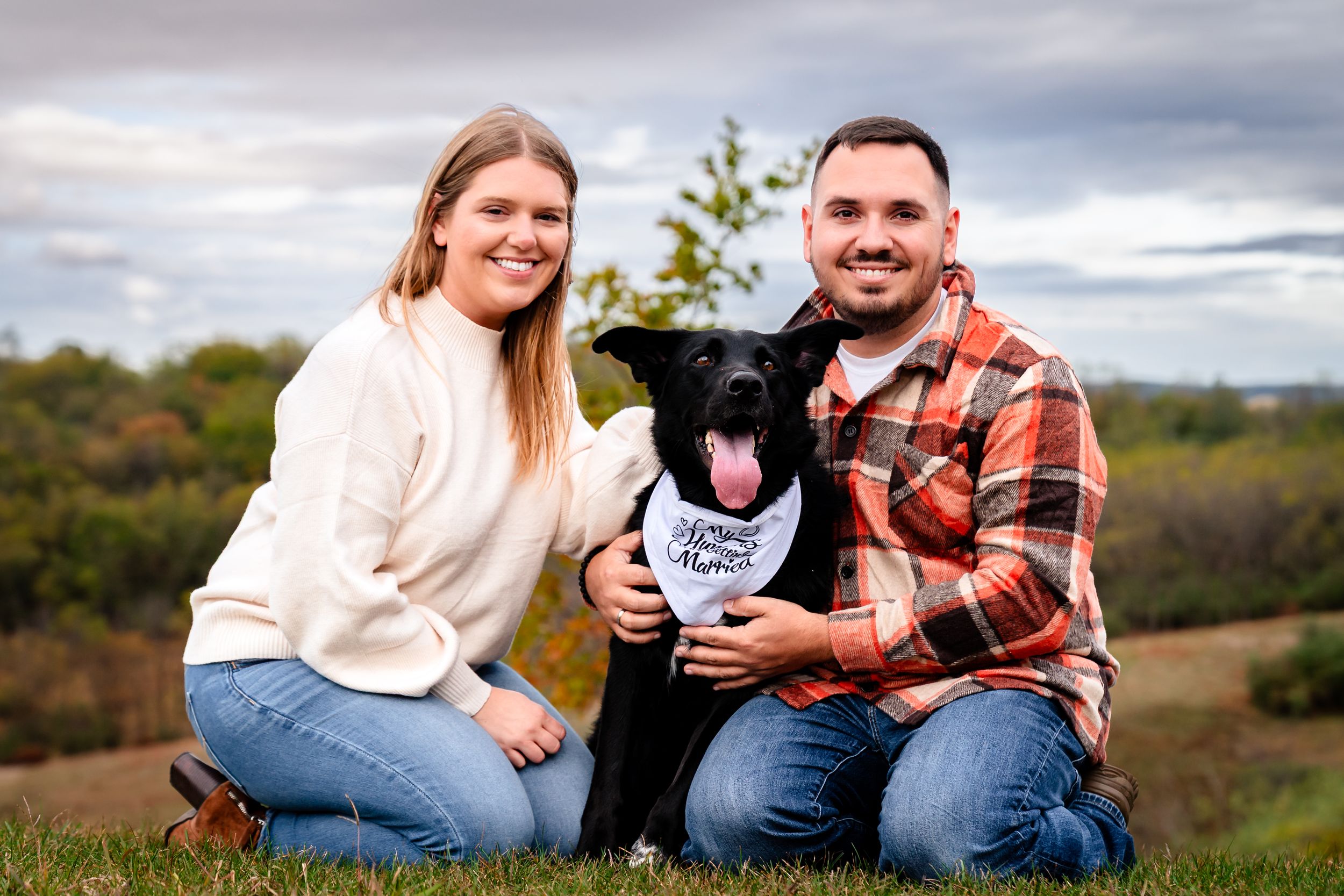 Couple poses with their black dog for fall family portrait on grassy hillside.