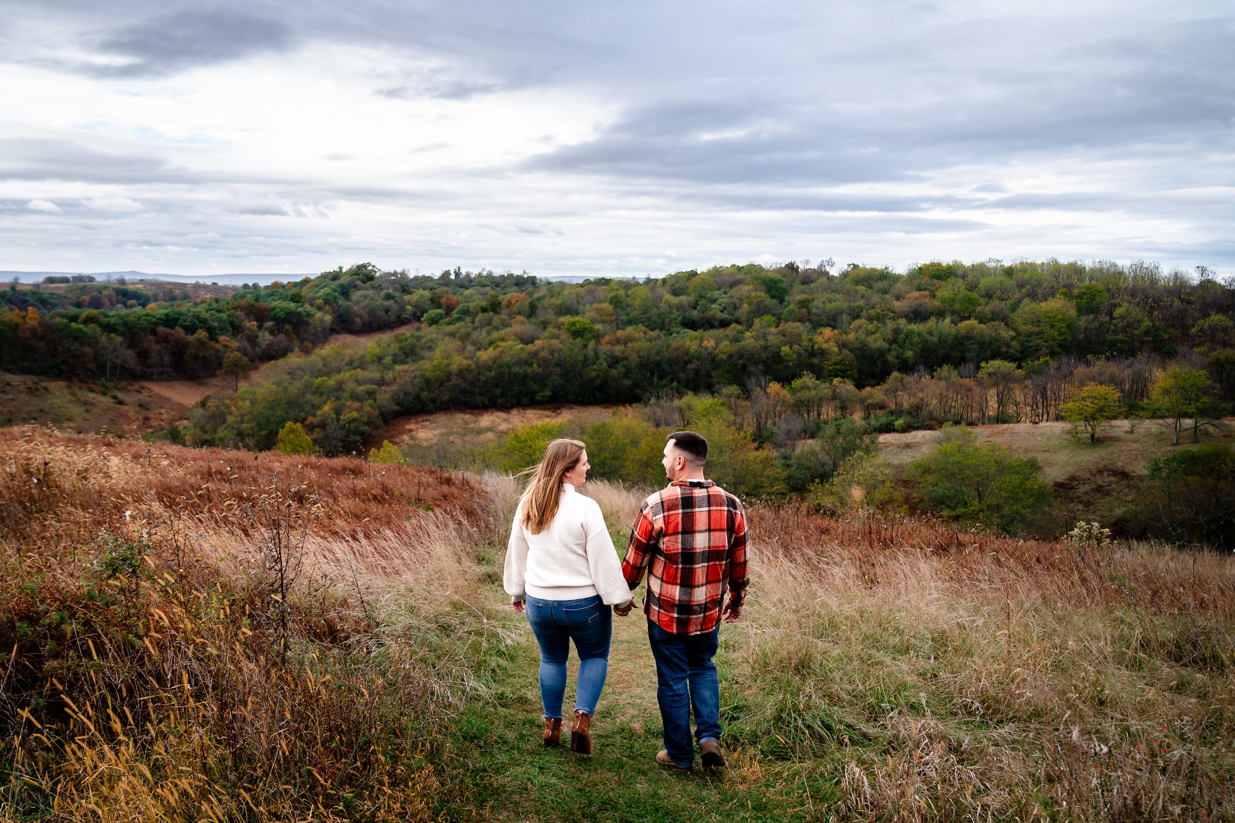 A couple walks through a scenic autumn field with rolling hills and cloudy skies in the background.