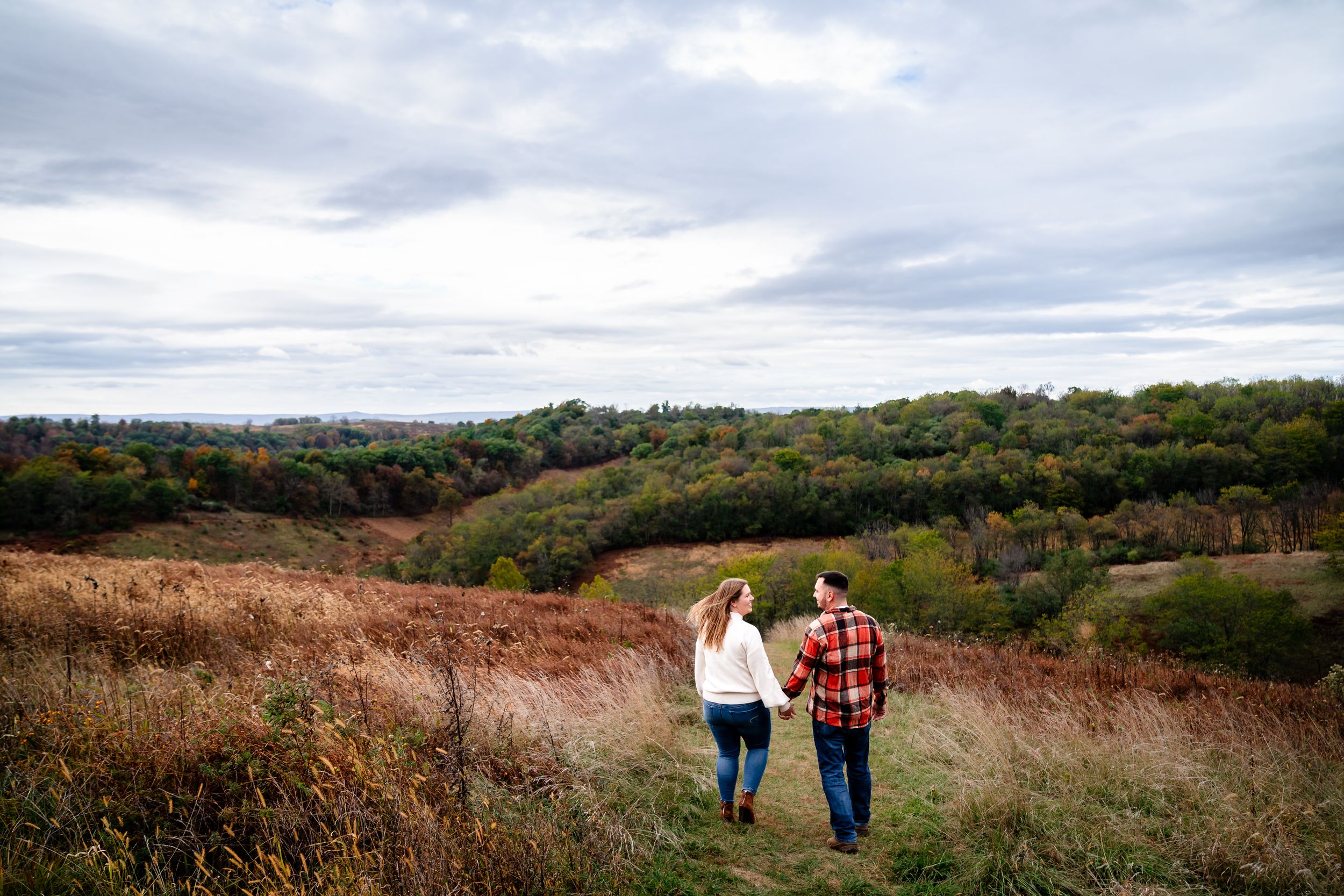 Two people stroll hand in hand through an autumn meadow with rolling hills in the distance.