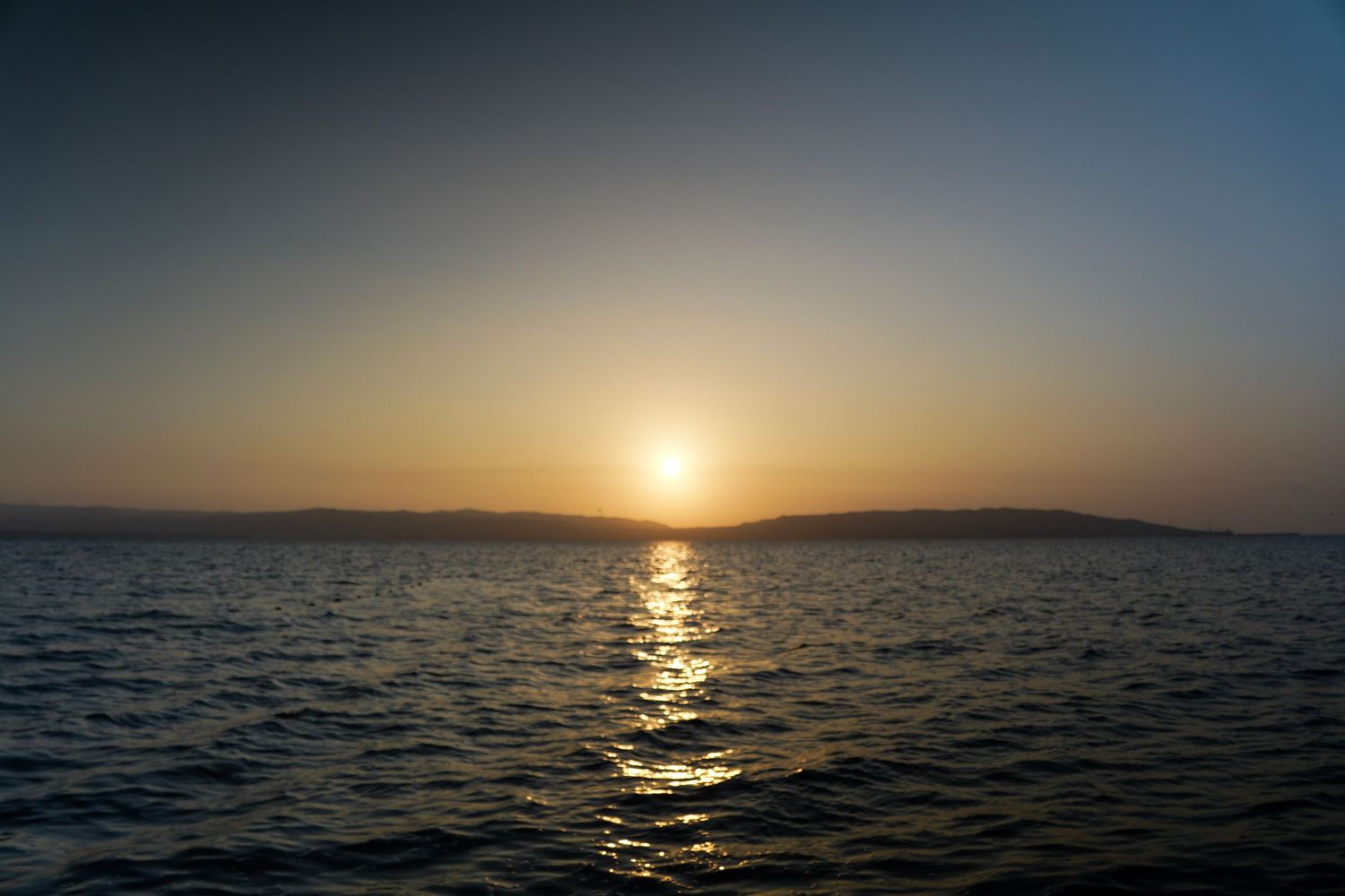 Golden sunset reflecting on calm ocean waters with distant mountain silhouettes on the horizon at dusk.