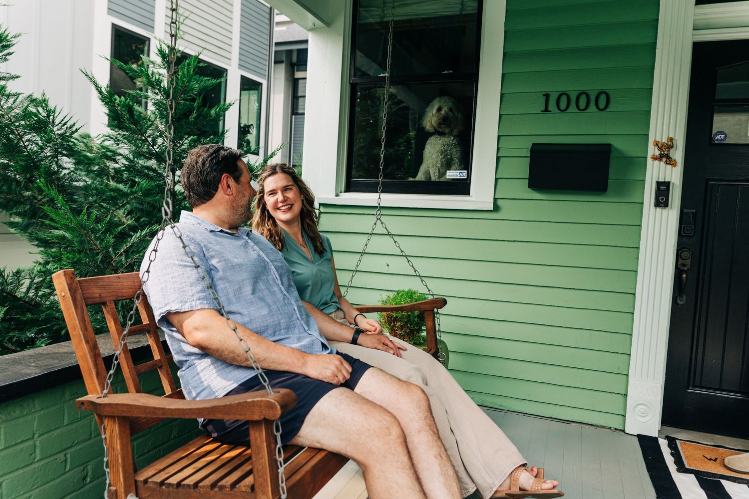 A couple relaxes together on a wooden bench on the porch of a mint green house in Charlotte.