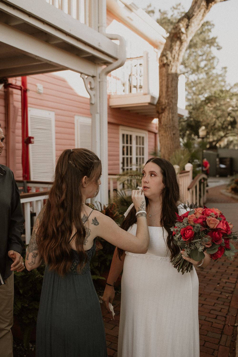 A wedding scene on a brick sidewalk outside a pink house with someone helping adjust makeup before the ceremony.