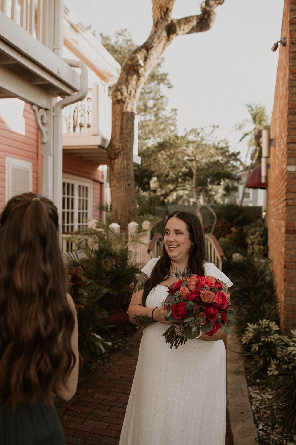 A bride in a white wedding dress holds a vibrant red bouquet while posing in front of a brick building at sunset.
