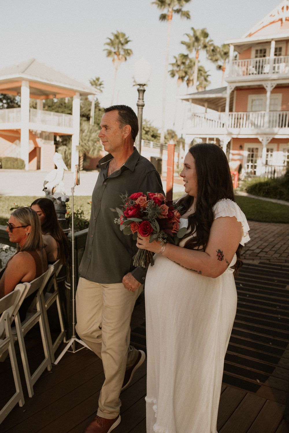 A couple walks down an outdoor aisle at a waterfront wedding ceremony with palm trees and white buildings in the background.