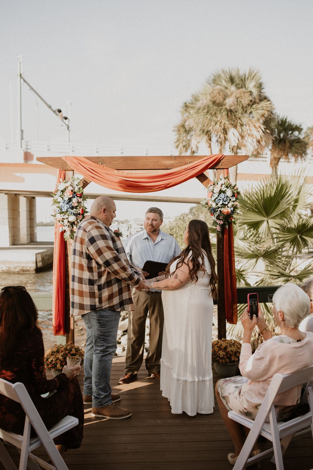 Intimate waterfront wedding ceremony with coral draping and floral arch at sunset.