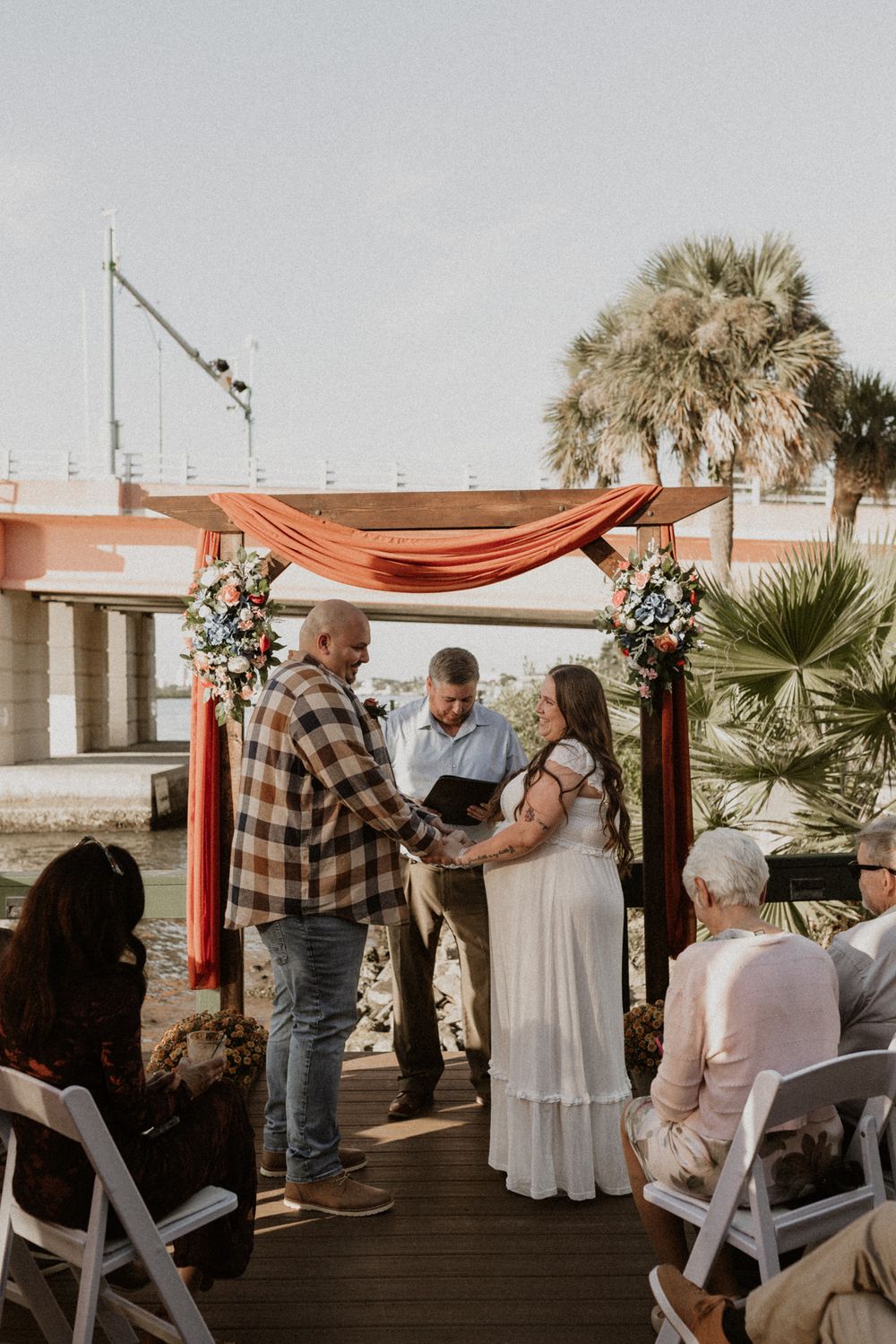 A romantic outdoor wedding ceremony setup with white chairs and coral drapes against a waterfront backdrop.