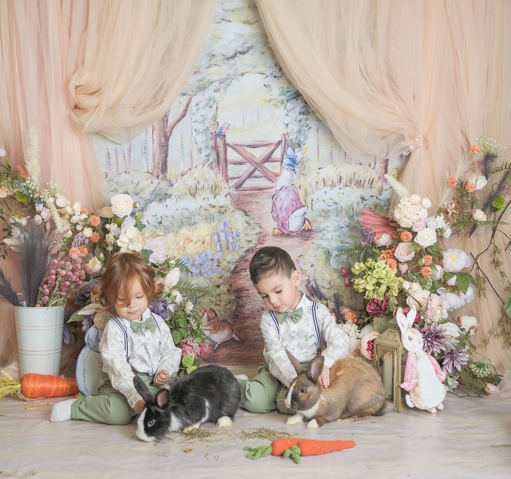 Two children sit on a floral set with bunnies and carrots in a dreamy spring-themed photography setup.