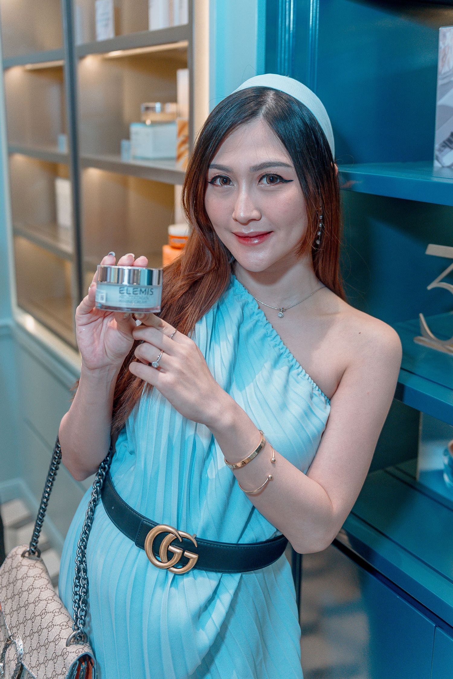 Woman in light blue pleated dress with Gucci belt holds small product while posing near blue shelving unit.