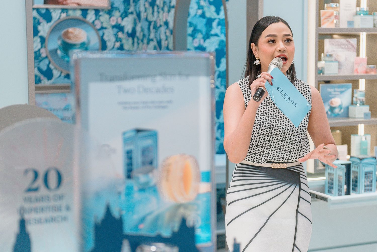 A woman in a polka dot top and striped skirt stands in front of a retail display with blue product packaging.